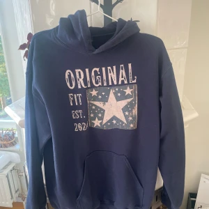 Mörkblå hoodie - Mörkblå hoodie från Original Fit med stor vit stjärna och text på bröstet. Tröjan har huva, magficka och är tillverkad i en mjuk bomulls- och polyestermix. Perfekt för en chill och avslappnad stil. Strl M