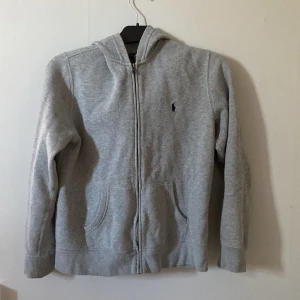 Grå hoodie från Polo Ralph Lauren - Klassisk grå hoodie från Polo Ralph Lauren med dragkedja framtill, huva och två fickor. Mjuk fleece på insidan och broderad logga på bröstet. Perfekt för en chill och avslappnad stil.