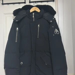 Svart dunjacka Moose Knuckles - Svart dunjacka från Moose Knuckles med pälskantad huva och flera silvriga dragkedjor. Jackan har broderad patch på insidan med hockeymotiv, samt logga på ärmen. Snygg och varm modell med lång ärm och coola detaljer.