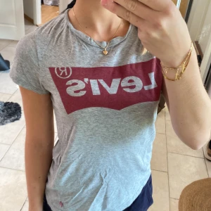 Grå Levi's t-shirt med röd logga - Säljer en grå t-shirt från Levi's i storlek XXS med den klassiska röda loggan tryckt framtill. T-shirten har korta ärmar och är gjord i mjuk bomull, perfekt för en chill och avslappnad stil. Passformen är normal och färgtonen är ljusgrå.