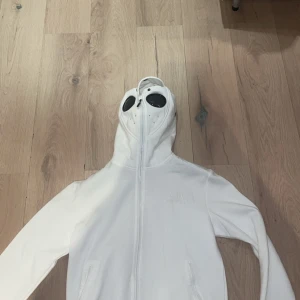 Vit hoodie med goggles från C.P. Company - Säljer en vit hoodie från C.P. Company med ikoniska goggles i huvan och dragkedja hela vägen upp. Tröjan har broderad logga på bröstet och två fickor framtill. Perfekt för dig som vill sticka ut med en unik streetwear-look.