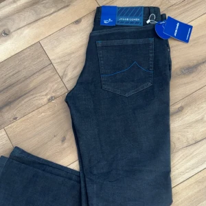 Jacob Cohën mörkblå jeans, stl 30 - Snygga mörkblå jeans från Jacob Cohën med klassisk femficksdesign och blå kontrastsömmar på bakfickorna. Jeansen har en rak passform och är tillverkade i mjukt denimtyg som sitter skönt hela dagen. Perfekta för dig som gillar stilrena och exklusiva jeans.