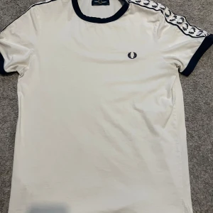 Vit Fred Perry t-shirt med logoband - Vit t-shirt från Fred Perry med marinblå detaljer runt hals och ärmslut. Ikoniskt Fred Perry-logoband längs axlarna och broderad logga på bröstet. 
