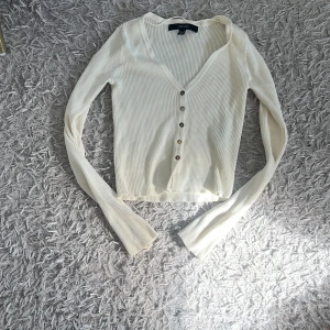 Vit ribbad croppad topp med knappar - Säljer en fräsch vit ribbad topp från Vero Moda med croppad passform och snygg v-ringning. Toppen har långa ärmar och fyra silvriga knappar framtill. Perfekt för dig som gillar stilrena och enkla plagg med lite edge.