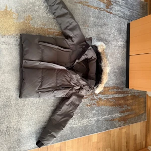 Grå Canada Goose dunjacka med pälskrage - Säljer en grå dunjacka från Canada Goose med snygg pälskant på huvan. Jackan har dragkedja, knappar och flera fickor framtill. Märkets klassiska patch syns på ärmen. Perfekt för kalla dagar och riktigt varm. Kommer med originalpåse.