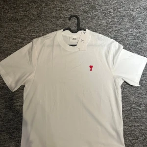 AMI vit T-shirt  - Vit t-shirt från Ami Alexandre Mattiussi med klassisk passform och korta ärmar. På bröstet finns ett broderat rött hjärta med bokstaven A. Tillverkad i mjuk bomull och har rund halsringning. Perfekt för en clean och stilren look.