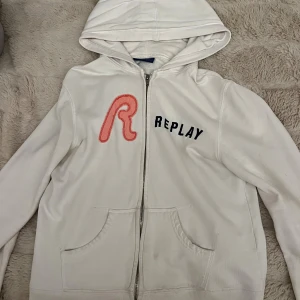 Vit hoodie med dragkedja från Replay - Vit hoodie från Replay med dragkedja framtill, stor röd broderad R och svart Replay-tryck på bröstet. Tröjan har huva, kängurufickor och är tillverkad i mjuk bomull. Perfekt för en avslappnad och cool stil. Tyvär är det lite sönder längst ner på ärmarna.