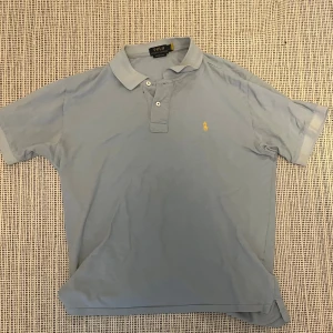Ljusblå pikétröja Polo Ralph Lauren L - Snygg ljusblå pikétröja från Polo Ralph Lauren med klassisk krage och två knappar framtill. Ikoniska gula loggan broderad på bröstet. Perfekt för en clean och stilren look.