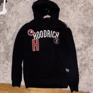Svart Hoodie från Hoodrich med patchar - Svart hoodie från Hoodrich med stor vit logga och röda samt svarta patchar framtill. Klassisk huva med snörning och känguruficka. Mjuk insida och ribbade muddar. Perfekt för dig som gillar streetwear och vill sticka ut med coola detaljer. Vill ni ha fler bilder hör av er! Pris kan diskuteras.