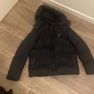 Svart pufferjacka Urban Classics - Svart pufferjacka från Urban Classics med huva och dragkedja framtill. Jackan är vadderad och har en stilren look med diskret logotyp på bröstet. Perfekt för kalla dagar när du vill hålla värmen och ändå se clean ut.