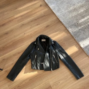 Svart skinnjacka från Bershka XS - Säljer en svart skinnjacka från Bershka i bikerstil med silvriga dragkedjor och detaljer. Jackan är croppad och har klassisk krage samt två fickor med dragkedja framtill. Perfekt för dig som vill ha en edgy look.
