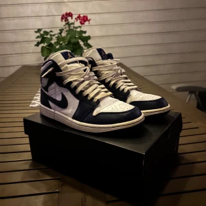 Nike Air Jordan 1 Mid Navy/Vit EU42 - Säljer ett par Nike Air Jordan 1 Mid i marinblått och vitt skinn. Klassisk high-top siluett med vit bas, mörkblå overlays och matchande Swoosh. Skorna har snörning och en rund tå. Perfekta för dig som gillar streetwear och sneakers med ikonisk design. Original kartong följer med och ett par nya mörkblå skosnören.