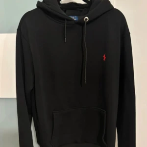 Svart hoodie från Polo Ralph Lauren - Svart hoodie från Polo Ralph Lauren med klassisk röd broderad logga på bröstet. Tröjan har huva med snörning, känguruficka och är långärmad. Perfekt för en chill och stilren look.