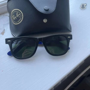 Ray-Ban Wayfarer svarta/blå solglasögon - Säljer ett par Ray-Ban Wayfarer solglasögon med klassisk svart front och coolt blått innerskaft med vit logga. Grönt glas och ikonisk form. Kommer med original Ray-Ban fodral. Perfekt för dig som vill sticka ut med stil.