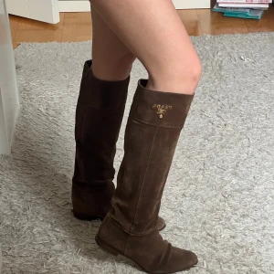 Bruna mockastövlar från Prada - Snygga knähöga stövlar från Prada i mörkbrun mocka med guldfärgad logga upptill. Perfekta  för höst och vinter! 