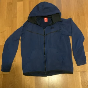 Blå hoodiejacka från Nike, storlek M - Blå hoodiejacka från Nike med hel dragkedja och svart Nike-logga på bröstet. Jackan har huva, två fickor fram och svarta detaljer längs dragkedjan och bröstet. Tillverkad i mjuk bomullsmix, perfekt för chill eller träning. Sitter mer som S