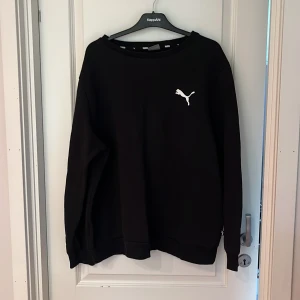 Svart sweatshirt från Puma XXL - Svart sweatshirt från Puma i storlek XXL. Tröjan har en klassisk rund hals, långa ärmar och en vit Puma-logga på bröstet. Tillverkad i mjuk bomullsmix som är skön att ha på sig. Perfekt för chill eller sportiga dagar.