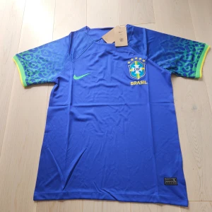 Blå Brasil Nike fotbollströja - Snygg blå Brasilien fotbollströja från Nike med CBF-emblem och gula detaljer. Ärmarna har leopardmönster i blått och grönt med gul kant. Tröjan är kortärmad och tillverkad i lätt, ventilerande polyester. Perfekt för dig som vill sticka ut på planen eller heja på Brasilien.