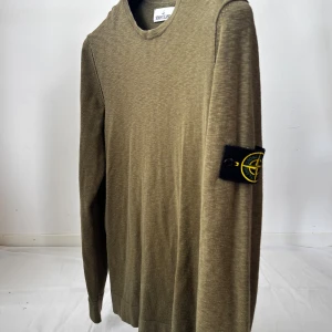 Olivgrön långärmad tröja Stone Island - Olivgrön långärmad tröja från Stone Island med klassisk rund hals och ikonisk logotyp-patch på vänster ärm. Tröjan är i mjuk bomull och har en clean, stilren look som passar perfekt till jeans eller cargobyxor.