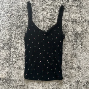 Svart blommig linnetopp med spets - Supersöt svart linnetopp med små broderade blommor i rosa och grönt. Toppen har spetskant längs urringningen och axelbanden, samt en liten rosett framtill. Perfekt för en trendig och feminin look.