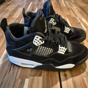 Svarta Nike Air Jordan 4 sneakers - Säljer ett par svarta Nike Air Jordan 4 sneakers med vita detaljer och meshpaneler på sidorna. Skorna har klassisk snörning, hög siluett och den ikoniska Jumpman-loggan på plösen och hälen. Ovandelen är i mocka och sulan är vit med svarta inslag.