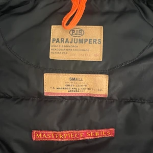 Svart Parajumpers pufferjacka med päls - Säljer en svart Parajumpers pufferjacka i storlek small. Jackan har en fluffig huva med äkta päls, coolt Parajumpers-märke på ärmen och orange detalj i nacken. Flera fickor framtill och dragkedja. Perfekt för kalla vinterdagar. äkta men finns en liten skada på luvan