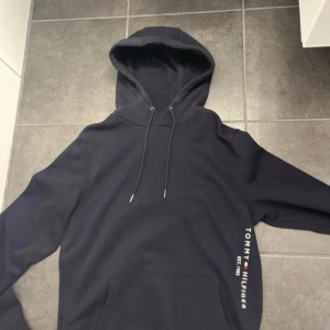 Mörkblå hoodie Tommy Hilfiger - Snygg mörkblå hoodie från Tommy Hilfiger med klassisk huva och dragsnören. Tryckt logga och text längs sidan framtill. Perfekt för dig som gillar stilrena och bekväma plagg. Stor känguruficka framtill och långa ärmar.