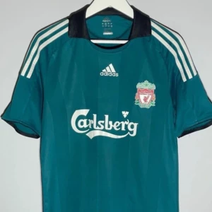 Liverpool Gerrard bortatröja Adidas - Liverpool FC bortatröja med Gerrard 8 på ryggen. Tröjan är mörkgrön med svarta och vita detaljer, Adidas-logga och Carlsberg-tryck på bröstet. Korta ärmar och klassisk passform, tillverkad i polyester. Perfekt för dig som älskar fotboll och retrostil.