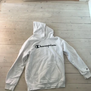 Vit hoodie från Champion - Säljer en vit hoodie från Champion med stor svart logga på bröstet och klassisk känguruficka. Tröjan har huva och långa ärmar med ribbade muddar. Perfekt för dig som gillar en clean och sportig stil.