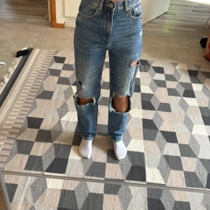 Blå mom jeans med slitningar från FB Sister - Säljer ett par blå mom jeans från FB Sister i storlek XXS. Byxorna har hög midja, raka ben och stora slitningar vid knäna för en cool, avslappnad look. Klassisk femficksmodell i jeansmaterial. Perfekta för dig som gillar en trendig och edgy stil.