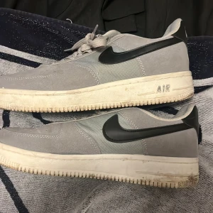 Nike Air Force 1 grå sneakers - Grå Nike Air Force 1 sneakers med svart swoosh och vit sula. Ovandelen är i mocka och textil, med klassisk snörning och perforerad tå för extra ventilation. Stilren och tidlös modell som passar till det mesta. Sulan har tydlig AIR-detalj på sidan.