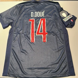 PSG matchtröja D. Doué #14 Nike - Paris Saint-Germain fotbollströja med D. Doué och nummer 14 på ryggen. Mörkblå med röd och vit detalj framtill, officiella UEFA Champions League och Foundation-märken på ärmen. Tillverkad i lätt och ventilerande polyester, klassisk passform. Replika, endast handtvätt gäller!