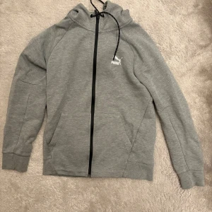 Grå hoodie från Puma med dragkedja - Snygg grå hoodie från Puma med svart dragkedja och huva med snörning. Tröjan har två fickor framtill och en stilren logga på bröstet. Perfekt för en avslappnad och sportig look.