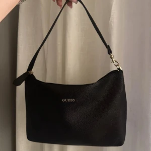 Svart axelväska från Guess i skinn - Stilren svart axelväska från Guess i mjukt skinn med guldfärgade detaljer och dragkedja upptill. Väskan har en enkel, klassisk form och logga framtill. Insidan är ljus med Guess-tryck. Perfekt storlek för det viktigaste.