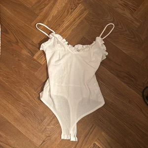Vit ribbad bodysuit med volang - Säljer en söt vit bodysuit med smala axelband och volangdetalj längs urringningen. Den är ribbad och har knäppning i grenen. Perfekt till jeans eller kjol för en fräsch look.