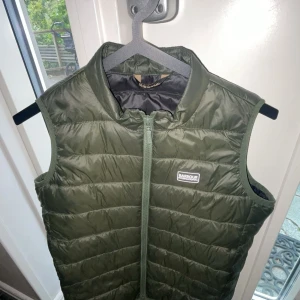 Olivgrön dunväst från Barbour XL - Snygg olivgrön dunväst från Barbour International i storlek XL. Västen har quiltad design, hög krage och dragkedja framtill. Perfekt att slänga över en hoodie för extra värme. Klassisk Barbour-logga på bröstet och lätt vadderad känsla.