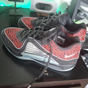 Nike KD 15 sneakers svart/röd - Nike KD 15 sneakers med svart och röd ovandel i mesh med mönstrad design, silverfärgade detaljer på sidorna och genomskinlig sula med orange inslag undertill. Skorna har snörning och låg profil, perfekt för dig som gillar sportig och modern stil. 