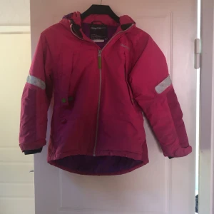 Vinterjacka från Didriksons - Säljer en rosa vindjacka från Didriksons med lila foder och huva. Jackan har reflexdetaljer på ärmar och rygg och dragkedja framtill. Storlek 120 och i fint skick