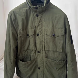 Olivgrön vindjacka Peak Performance XL - Olivgrön vindjacka från Peak Performance i storlek XL. Jackan har fyra stora fickor framtill, hög krage, knappar och dragkedja. Tillverkad i polyester och har en avtagbar huva. Perfekt för dig som gillar en stilren och funktionell look. (Skada på axeln)