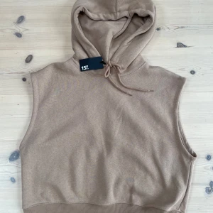 Beige hoodie-väst från 157 - Beige ärmlös hoodie-väst från 157 med huva och dragsko. Västen har ribbad nederkant och är tillverkad i mjukt bomullsmaterial. Perfekt för lager-på-lager och chill vardagsstil.