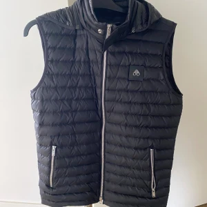 Svart dunväst Moose Knuckles med huva - Svart dunväst från Moose Knuckles med quiltad design och avtagbar huva. Västen har två fickor med dragkedja och en diskret logga på bröstet. Perfekt för lager-på-lager och snygg till streetwear. Lätt och smidig att bära.