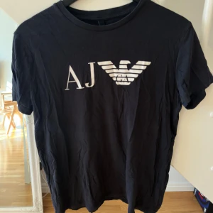 Svart t-shirt från Armani  - Svart t-shirt från Armani med stort vitt AJ-logotryck på bröstet. Tillverkad i 100% pima bomull för en mjuk och skön känsla. Klassisk rund halsringning och korta ärmar. Perfekt för en stilren och enkel look.