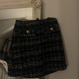 Svartvit rutig kortkjol med guldknappar - Elegant svart tweedkjol med vit rutstruktur och fina guldknappar framtill. Endast använd 1–2 gånger, i nyskick.