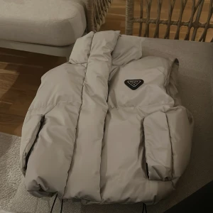 Beige dunväst från Prada - Säljer en beige dunväst från Prada med hög krage och ikonisk logga på bröstet. Västen har dragkedja och tryckknappar framtill samt två sidofickor med dragkedja. Den är puffig och har en clean, minimalistisk look som är riktigt snygg till streetwear.