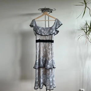 Blå silvrig off-shoulder spetsklänning med volang - Romantisk blåsilvrig off-shoulder spetsklänning med blommigt mönster och volangdetaljer längs axlar och nederkant. Perfekt för fest, finare tillställning, sittning mm. 