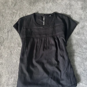 Svart blus med spetsdetaljer XS - Svart blus från H&M i storlek XS. Blusen har korta ärmar och fina spetsdetaljer längs halsringning och ärmslut. Den har dekorativa sömmar och lätt rynkade partier framtill. Knäppning med knapp i nacken. 