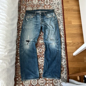 Levi's 501 slitna blå jeans - Säljer ett par vintage Levi's 501 jeans i blå tvätt med slitningar och hål på benen för en riktigt rå streetstil. Jeansen har raka ben, normal passform och snyggt slitna detaljer. Perfekta för dig som gillar vintagevibe och avslappnad look. Jeansen är 100 cm lång o 40 i midjan