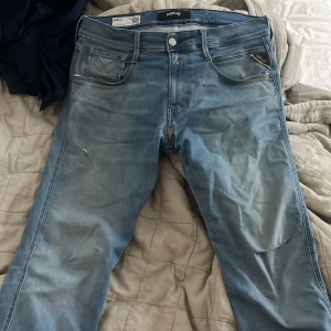 Replay Anbass ljusblå jeans slim fit - Snygga ljusblå Replay Anbass jeans i slim fit med klassisk femficksdesign och diskreta slitningar. Jeansen har en modern passform, normal midja och är tillverkade i mjukt stretchigt denim. Ett hål vid knät på ena sidan annars inga större defekter storlek 32 W 30 L. Går att pruta priset vid smidig affär.