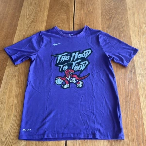 Lila Nike Dri-Fit t-shirt med tryck Toronto Raptors - Lila Nike Dri-Fit t-shirt med coolt tryck på bröstet: 'The Need To Feed' och en tecknad röd dinosaurie med basketboll. Klassisk rund hals och korta ärmar. Perfekt för dig som gillar sportig streetstyle och vill sticka ut.