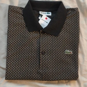 Mönstrad brun Lacoste Piké  - Snygg svart pikétröja från Lacoste med små geometriska mönster i beige. Klassisk krage, korta ärmar och knappar framtill. Ikoniska krokodilloggan broderad på bröstet. Tillverkad i 100% bomull i storlek M. Se mått i bilderna📏 Oanvänd i nyskick⭐